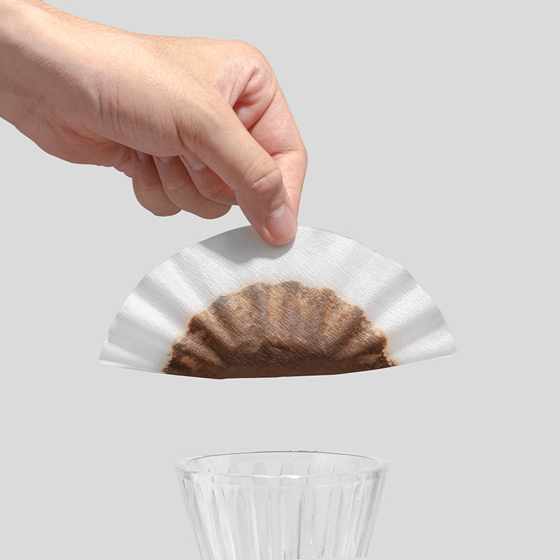 Hand holding a coffee filter with coffee grounds above a clear glass on a light gray background